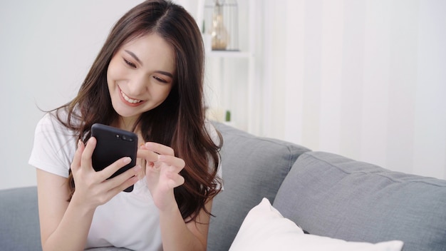 Asian woman playing smartphone while lying on home sofa in her living room.