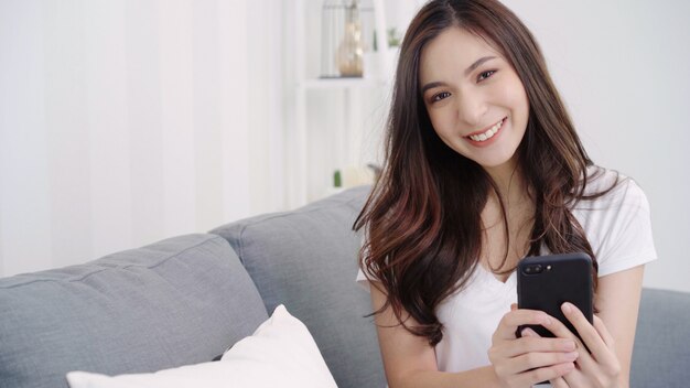 Asian woman playing smartphone while lying on home sofa in her living room.