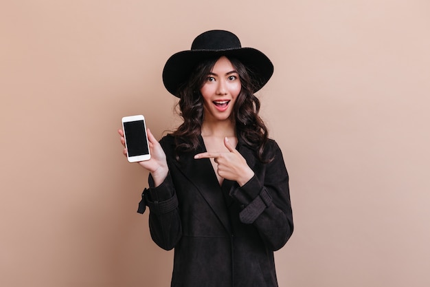 Азиатская женщина указывая пальцем на смартфон с пустым экраном. Студия выстрел эмоциональной корейской леди в черном пальто.