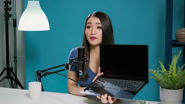 Asian woman reviewing laptop computer on social media podcast, recording live network video. Female vlogger broadcasting product recommendation with modern gadget and online technology.