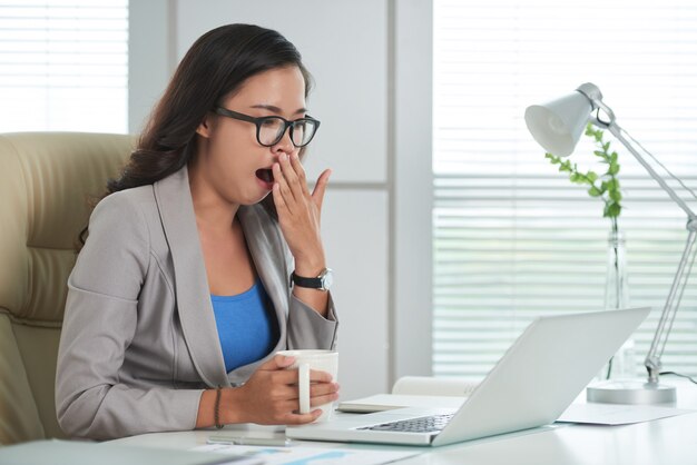 Asian woman sitting at desk in office, looking at laptop screen and yawning