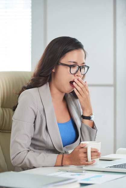 Asian woman sitting at desk in office with mug and yawning