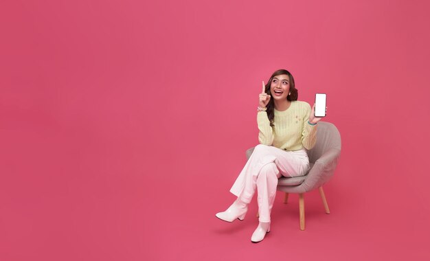 ソファに座って幸せを感じ、モバイルスマートフォンの空白の画面を示すジェスチャーをするアジアの女性