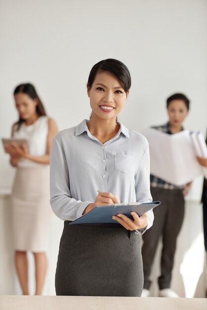 Asian woman in smart clothes posing in studio with clipboard, and colleagues in background