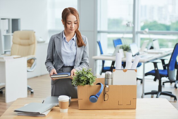 Asian woman standing at deks in office with belongings in cardboard box