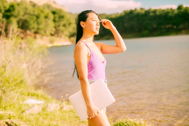 Asian woman standing near lake and holding laptop