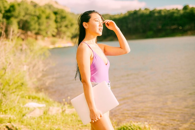 アジアの女性の湖の近くに立っているとラップトップを保持