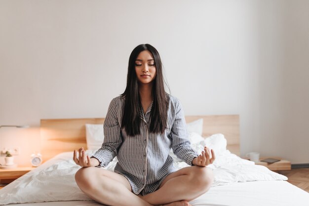 Asian woman in summer pajamas meditates sitting on white bed