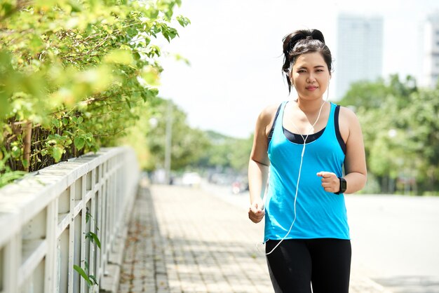 Asian woman in tank top and leggings, with earphones, jogging in morning in urban street 