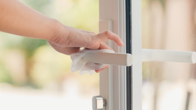 Asian woman using alcohol spray on tissue clean doorknob before open door for protect coronavirus. Female clean surface for hygiene when social distancing stay at home and self quarantine time.