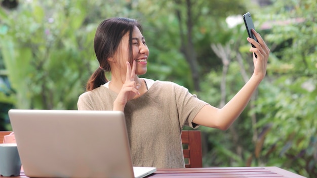 Asian woman using mobile phone selfie post in social media, female relax feeling happy showing shopping bags sitting on table in the garden in morning.