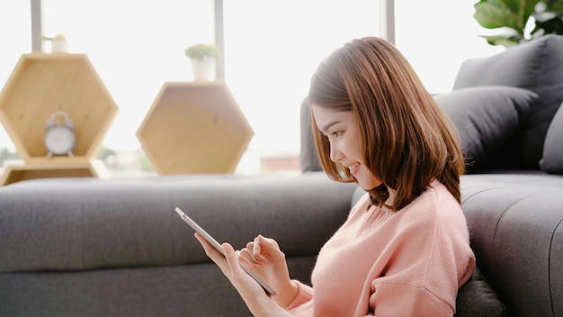 Asian woman using tablet while lying on home sofa in her living room. 