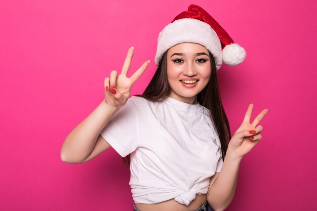 Asian woman wearing Santa's hat with peace gesture isolated on pink wall
