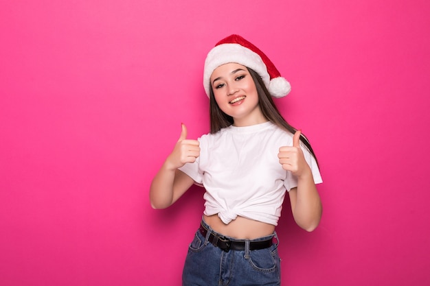 Азиатская женщина в шляпе Санта-Клауса с большими пальцами руки вверх изолирована на розовой стене