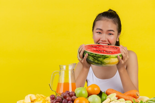 An Asian woman wearing a white tank top. Both hands hold watermelons and the table is full of various fruits.