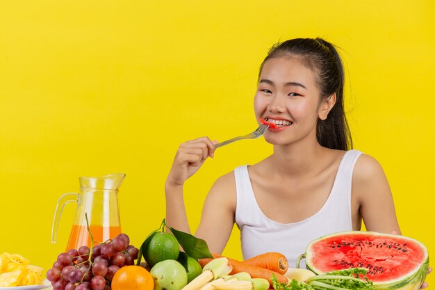 An Asian woman wearing a white tank top eating watermelon and the table is full of various fruits.