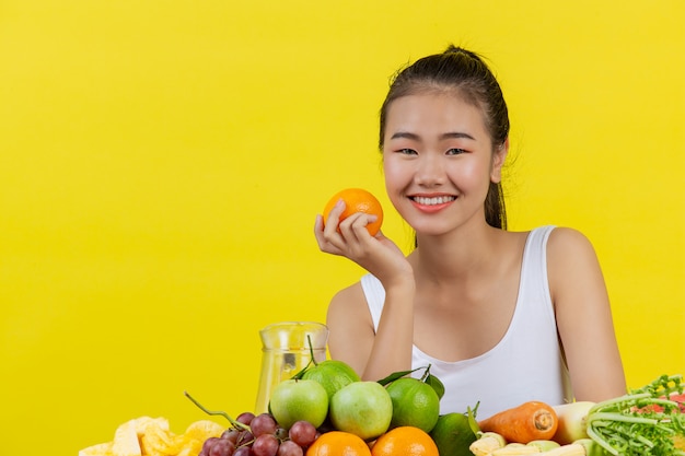 白いタンクトップを着ているアジアの女性。オレンジを右手で持ち、テーブルにはたくさんの果物があります。