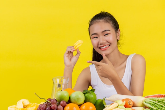 An asian woman wearing a white tank top. holding pineapples with his right hand and on the table there are many fruits