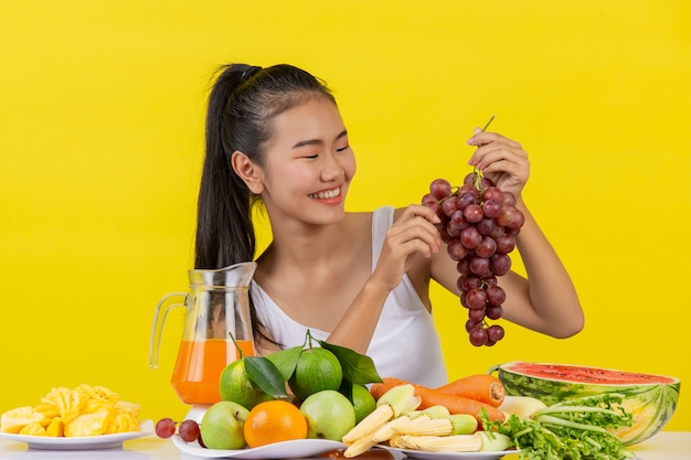 An asian woman wearing a white tank top. the left hand holds a bunch of grapes. the right hand picks up the grapes to eat and the table is full of various fruits