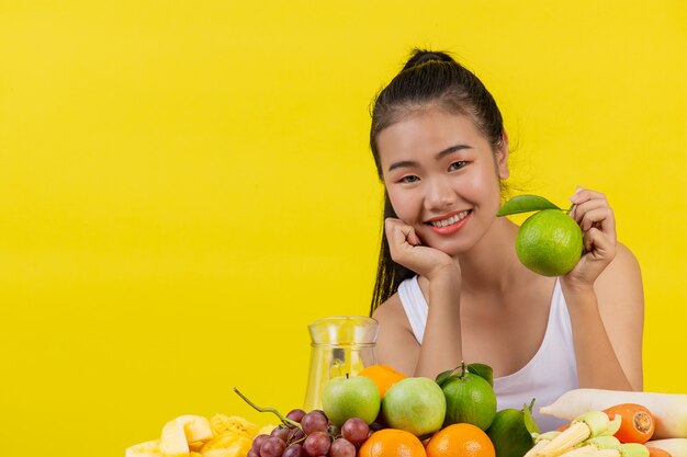An Asian woman wearing a white tank top. The left hand holds a green oranges and the table is full of many kinds of fruits.