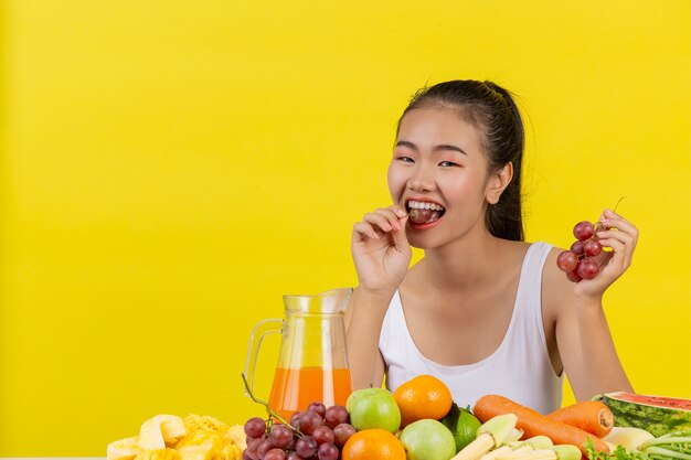 An Asian woman wearing a white tank top. The right hand holds a bunch of grapes and the table is full of various fruits.