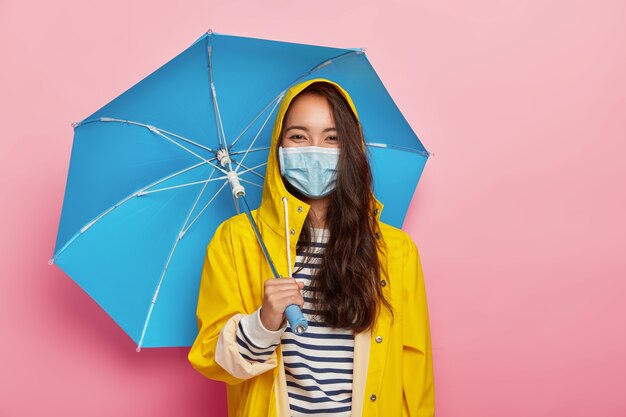 Asian woman wears protective mask, faces air pollution during rainy day, stands under umbrella, dressed in yellow raincoat