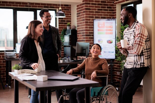 Asian woman in wheelchair and group of people laughing on job break, having fun in disability friendly office. Worker with chronic health condition chatting with cheerful colleagues.