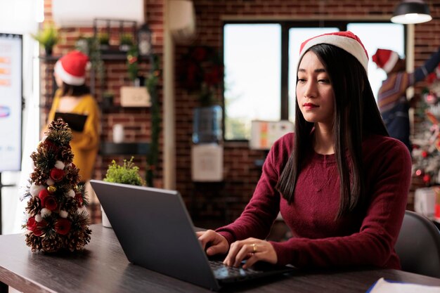 장식된 사무실에서 노트북 작업을 하는 아시아 여성, 비즈니스 보고서를 사용하여 스타트업 회사에서 일합니다. 직장에서 크리스마스 장식으로 크리스마스 트리와 축제 장식품을 즐깁니다.