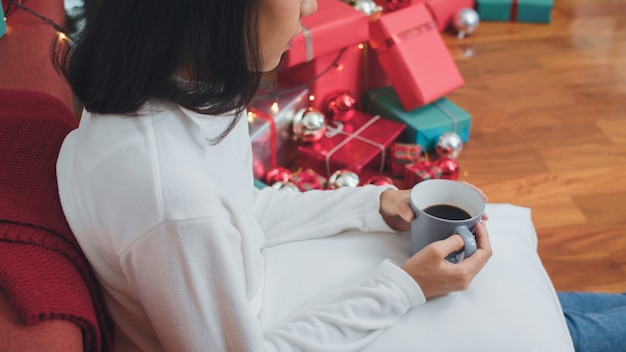 Asian women celebrate Christmas festival. Female teen relax happy having breakfast drink coffee near Christmas tree lying on sofa enjoy xmas winter holidays in living room at home.