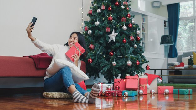 아시아 여성들은 크리스마스 축제를 축하합니다. 여성 청소년 행복 들고 선물을 휴식 하 고 크리스마스 트리와 스마트 폰 selfie를 사용 하여 집에서 거실에서 크리스마스 겨울 휴가 즐길 수 있습니다.