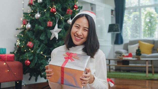 Asian women celebrate Christmas festival. Female teen wear sweater and Christmas hat relax happy hold gift smiling near Christmas tree enjoy xmas winter holidays together in living room at home.