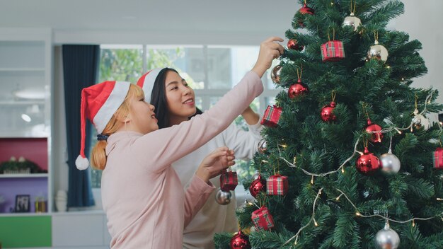 Asian women friends decorate Christmas tree at Christmas festival. Female teen happy smiling celebrate xmas winter holidays together in living room at home.