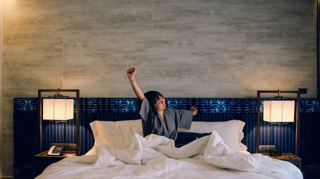 無料写真 アジアの女性は眠りから目覚める。週末の午前中にストレッチして、リラックスした週末のコンセプトの豪華な部屋のベッドに座っています