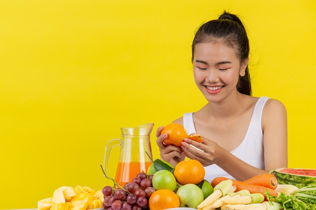 Asian women wear a white tank top. Holdingorange and a table full of many fruits.