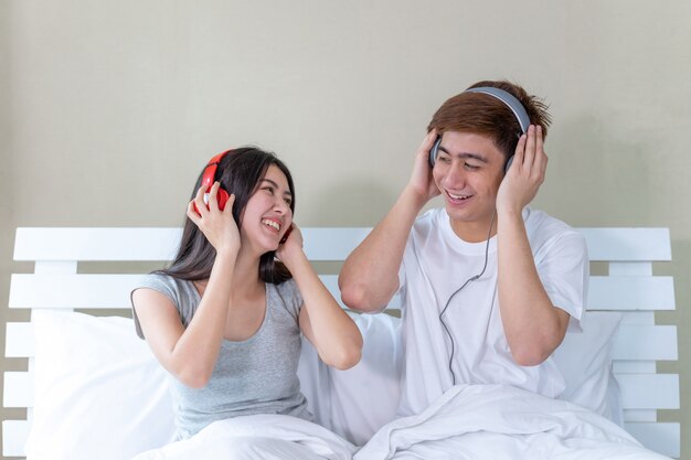 アジアの若いカップルがベッドの上に座って、ヘッドフォンで音楽を聴くとダンスを楽しく
