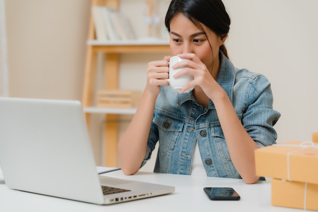 Giovane proprietario di donna asiatica di affari dell'imprenditore del prodotto in linea di controllo delle pmi sulle azione risparmia al computer e bevendo caffè che lavora a casa.