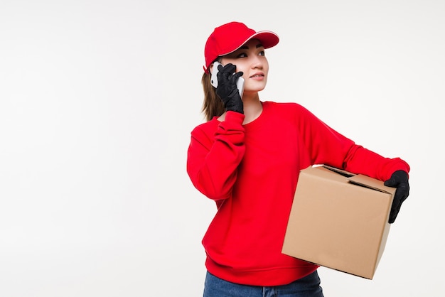 Asian young female delivery worker standing in white wall using mobile smartphone talking with customer checking shipping address