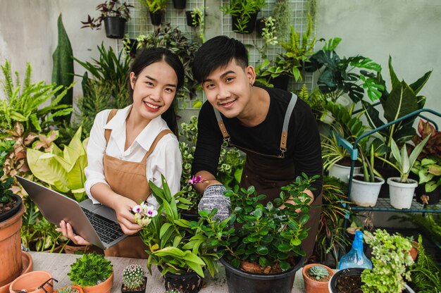 エプロンを身に着けているアジアの若い庭師のカップルは、温室内の観葉植物を研究し、世話をするために庭の機器とラップトップコンピューターを使用しています