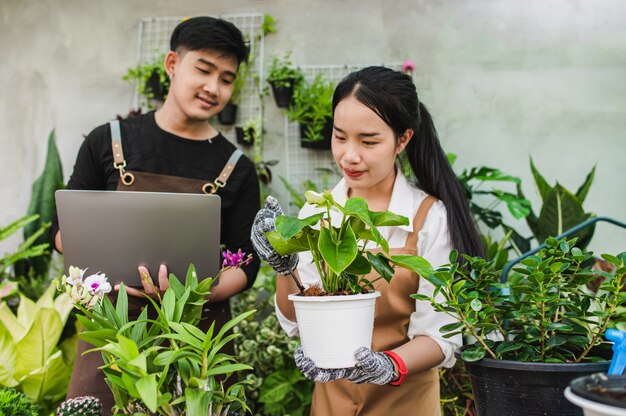 エプロンを身に着けているアジアの若い庭師のカップルは、温室内の観葉植物を研究し、世話をするために庭の機器とラップトップコンピューターを使用しています