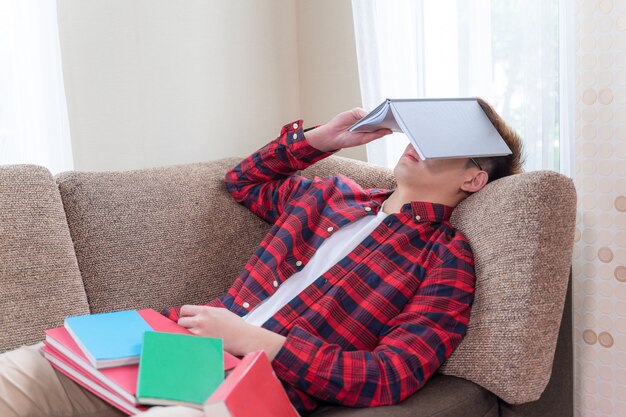 Азиатский молодой человек спит на диване, а стопка книг на его теле