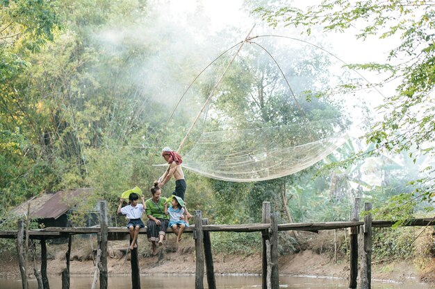 Asian young mother sitting on wooden bridge enjoy to play with two little daughters in student uniform hold banana leaf in hand, senior man walk with fishing net, copy space, rural lifestyle concept