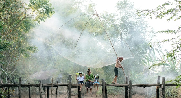 Asian young mother sitting on wooden bridge enjoy to play with two little daughters in student uniform hold banana leaf in hand, senior man walk with fishing net, copy space, rural lifestyle concept