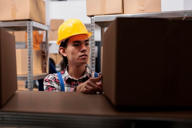 Asian young postal worker doing parcels organization in warehouse. Storehouse order picker in yellow hardhat looking at packages on shelf and searching for cardboard box close up