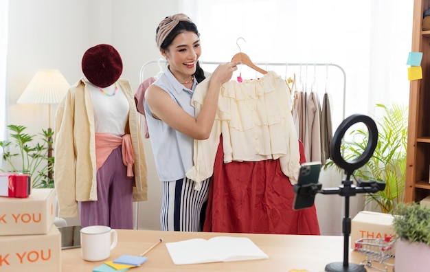 Asian young woman blogger showing clothes in front of smartphone camera while recording vlog video and live streaming at her shop SME or freelance business online and delivery concept