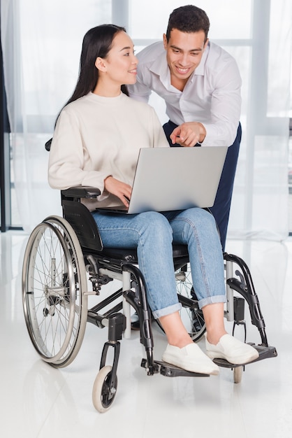 Asian young woman sitting on wheel chair looking at man showing something on laptop
