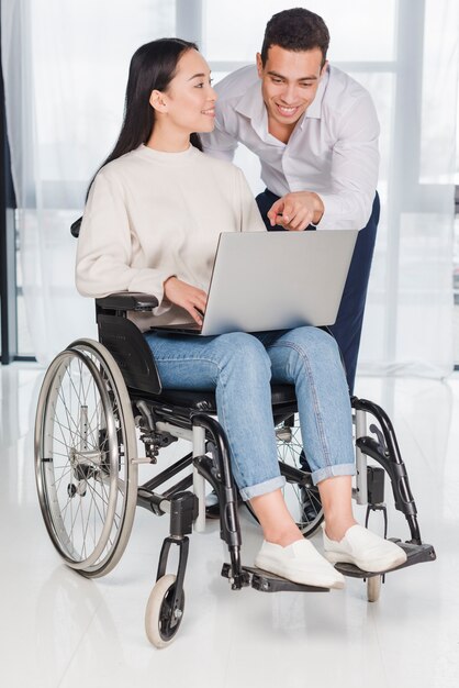 Азиатская молодая женщина, сидя на инвалидной коляске, глядя на человека, показывая что-то на ноутбуке