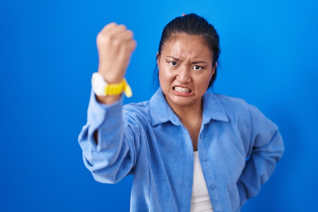 Free photo asian young woman standing over blue background angry and mad raising fist frustrated and furious while shouting with anger rage and aggressive concept