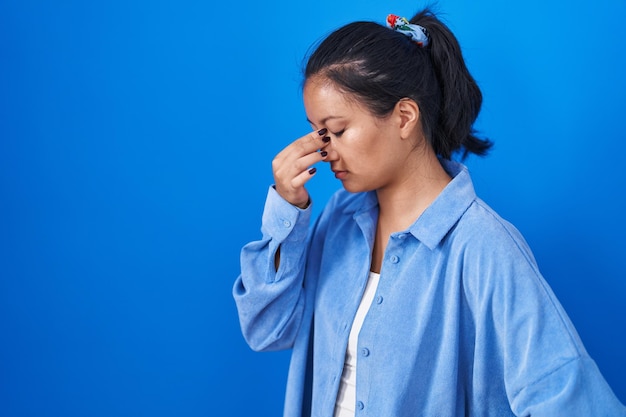 파란색 배경 위에 서 있는 아시아 젊은 여성은 피로와 두통을 느끼며 코와 눈을 문지르고 있습니다. 스트레스와 좌절 개념.