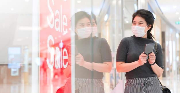 アジアの若い女性がフェイスマスク保護を身に着けているハンドホールドスマートフォン立っている店の看板ショーの販売促進を見てcovid19が広がった後の新しい通常のショッピングライフスタイル