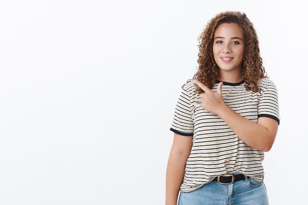 Assertive confident attractive chubby curly-haired girl pointing left index fingers standing aside showing white copy space